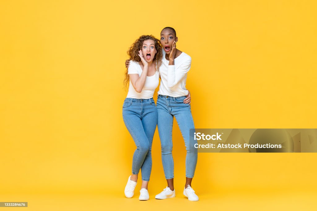 Full length portrait of shocked interracial millennial woman friends gasping with hands cupping mouths in isolated studio yellow color background Jeans Stock Photo