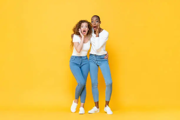 Photo of Full length portrait of shocked interracial millennial woman friends gasping with hands cupping mouths in isolated studio yellow color background