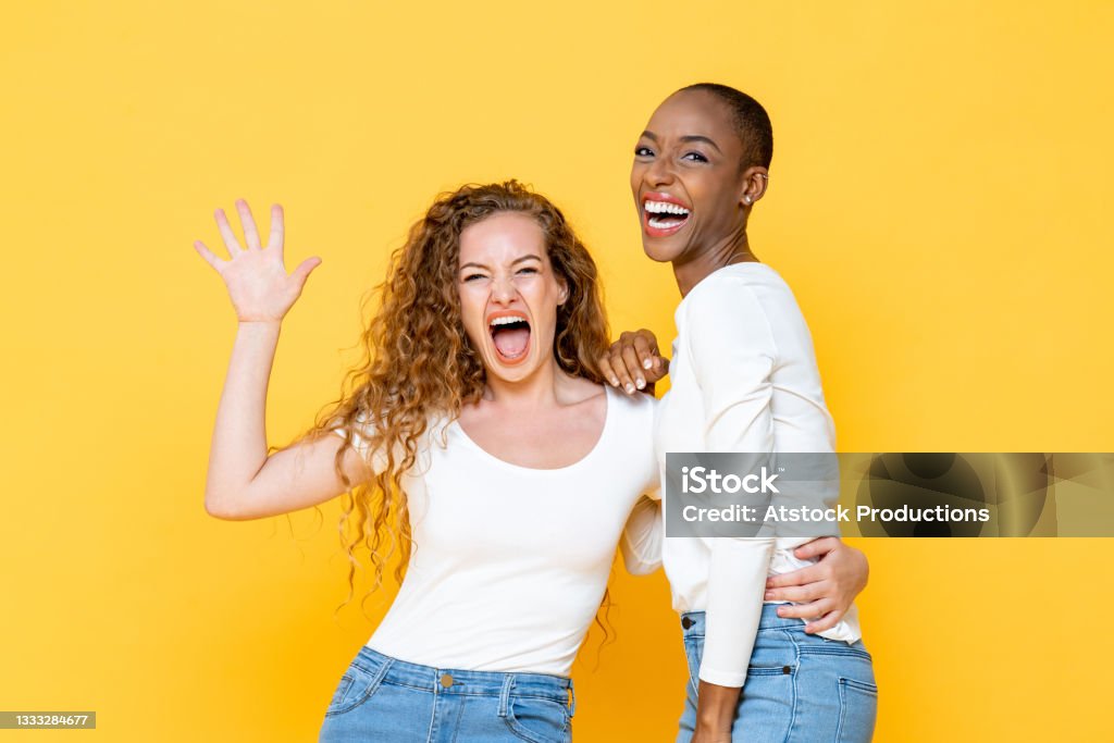 Portrait of surprised joyful interracial woman friends looking at camera on isolated yellow studio background Friendship Stock Photo