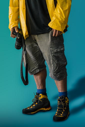 Hispanic adult male photographer wearing rain cover and hiking shoes while carrying his reflex camera against a blue background. Costa Rica, Centro America.