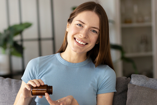 Millennial generation beautiful woman holding glass of water and food supplement capsule or antibiotic antidepressant. Looking at the camera and smiling