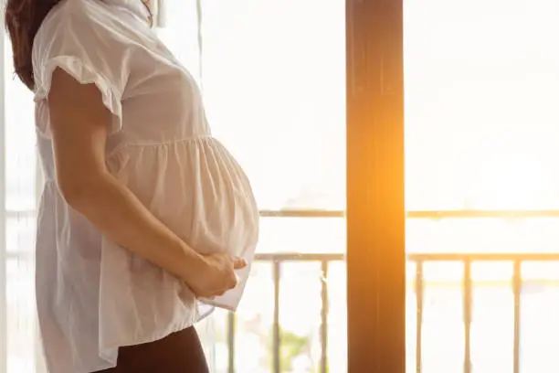 Photo of pregnant woman is enjoying herself waiting for her delivery and is relaxing on the white sofa by the window. Happy concept of pregnant women and looking forward to the day seeing the baby face.