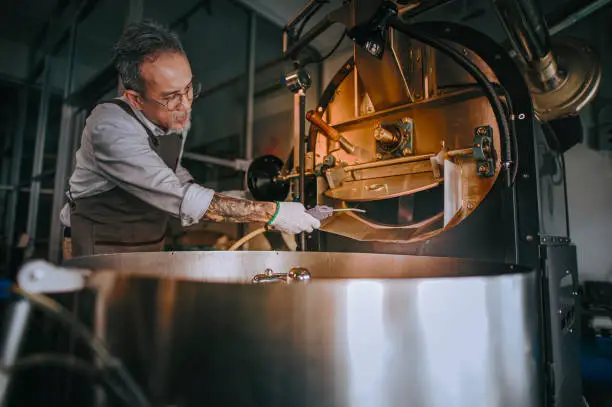 Photo of Asian chinese working senior man with glove brushing and cleaning coffee roaster machine with vacuum cleaning