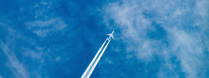 Distant passenger jet plane flying on high altitude through white clouds on blue sky leaving white smoke trace of contrail behind. Air transportation concept