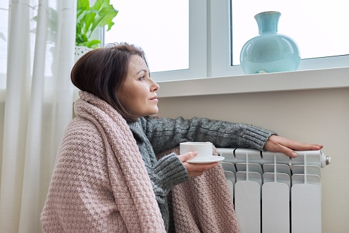 Winter season, woman warming up near home heating radiator. Female in warm woolen sweater under blanket with cup of hot drink