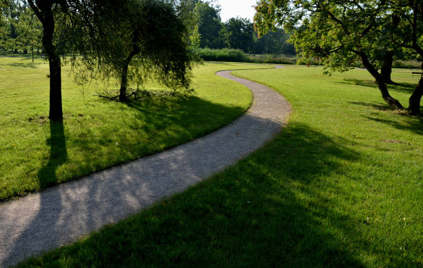 caminho do parque empoeirado ao longo da lagoa sinuoso através de um prado. leva à ponte e a parte do caminho são bancos longos com tábuas de madeira e plantas ornamentais vermelhas, brancas, perenais - road long dirt footpath - fotografias e filmes do acervo