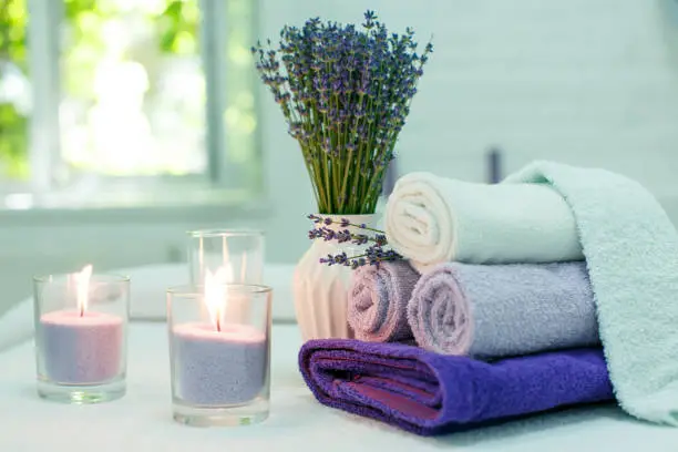 Photo of Three lit candles with loose wax, rolled lavender colored towels and a bouquet of blooming lavender on a couch in a massage parlor