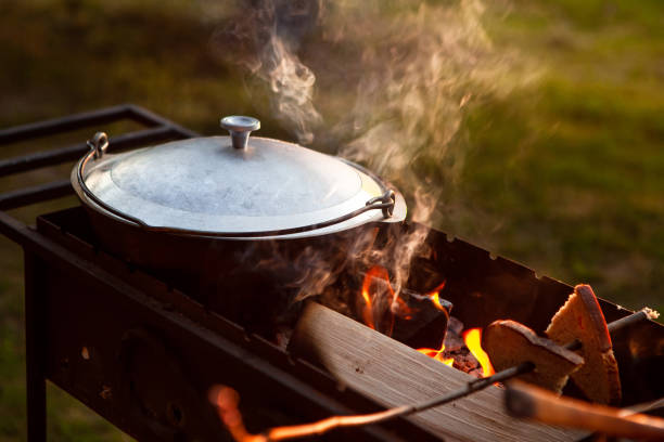 A saucepan, cauldron closed with a lid, in a grill on fire, from which steam comes out, on a natural background. Cooking in a pot on the fire. Camping concept.