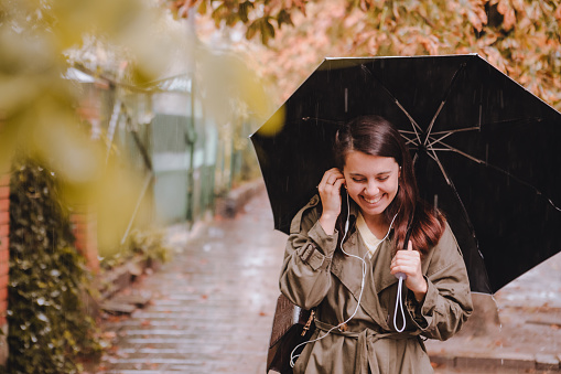 young pretty woman portrait listening music with headset while walking by street with umbrella under rain. copy space