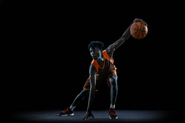 istock Vertical portrait. Young african man, basketball player practicing with ball isolated over dark studio background. 1333346380