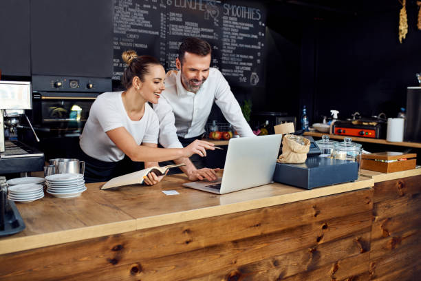 dos propietarios de cafeterías trabajando juntos planeando pedidos de suministro en una computadora portátil - small business owner fotografías e imágenes de stock