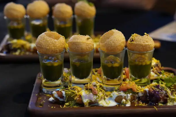 Indian chaat snack pani puri or golgappa served in shot glasses with tikki, curd, chutney, sev on brown rectangle wooden plate