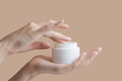 Women's fingers collect cream from round white jar. Concept of mockup for cosmetic products. Close-up, beige isolated background, copy space.