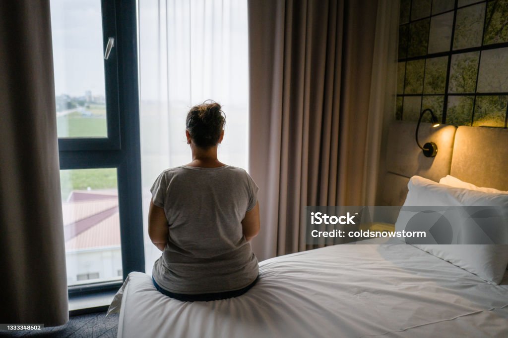 Woman sitting alone on bed in hotel room Color image depicting the rear view of a mid adult woman sitting on the edge of her hotel room bed. Abuse Stock Photo