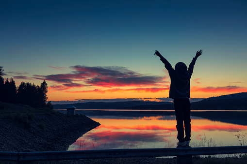 A teenage girl standing on a barrier at the shore of a calm lake at night with arms outstretched, enjoying the vividly colored evening in nature. Älvdalen, Sweden.