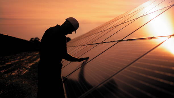 assistance technical worker in uniform is checking an operation and efficiency performance of photovoltaic solar panels. unidentified solar power engineer touches solar panels with his hand at sunset - solar panel fotos imagens e fotografias de stock
