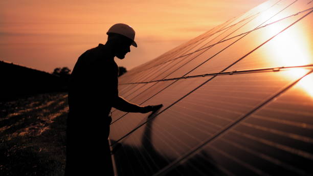 l'addetto tecnico dell'assistenza in uniforme sta verificando un funzionamento e le prestazioni di efficienza dei pannelli solari fotovoltaici. l'ingegnere dell'energia solare non identificato tocca i pannelli solari con la mano al tramonto - renewable energy photography color image horizontal foto e immagini stock