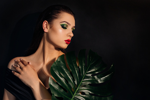 Close up face of beautiful young woman covering by green monstera green palm. Portrait of beauty woman with makeup red lips clear skin with big green glossy leaves.