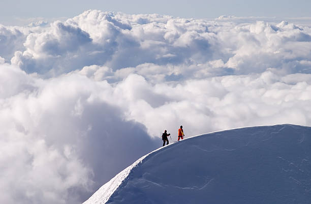 Mountaineers returning from the summit of Mont Blanc, late in the afternoon, passing a crest and heading the final few hundred meters to their shelter - Goûter Hut. The sky is clear, the weather is sunny, the team is walking above picturesque clouds.