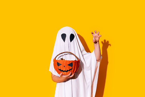Child in ghost costume holding basket of chocolates in one hand, and raised the other hand up.