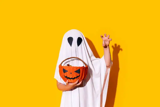 Photo of Child in ghost costume holding basket of chocolates in one hand, and raised the other hand up