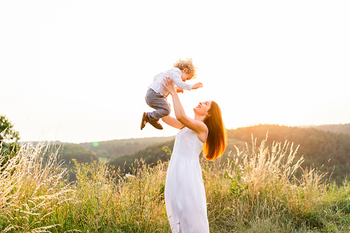 Happy mother with her son having fun in the field at sunset. Woman in a summer dress throws the child up