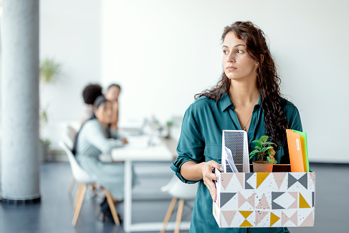 Portrait of young beautiful woman employee getting fired from work. Female walks through the office, carrying box with personal belongings. Business, firing and job loss concept