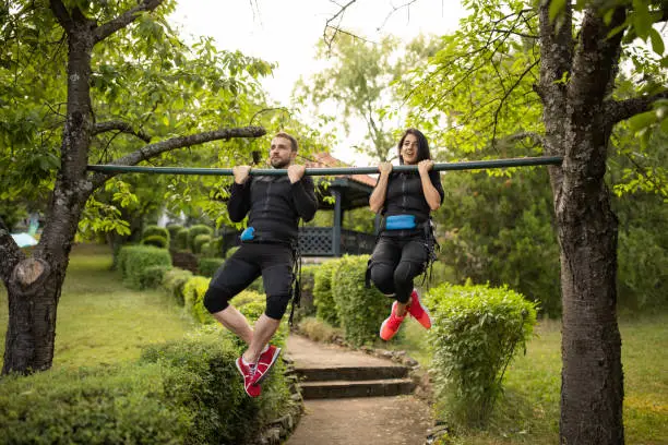 Young couple doing chin-ups while wearing electro muscle stimulation suit outdoors