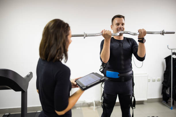 istock Young man exercising with barbell while wearing electro muscle stimulation suit in exercise room with fitness trainer beside him 1333368462