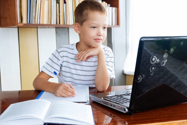 istock Distance learning online education. Caucasian schoolboy boy studies at home and does school homework, writes in workbook and looks at laptop 1333373073