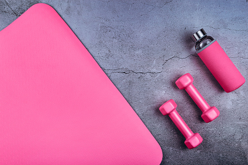 Top view of sport mat, glass water bottle and pink dumbbells on marble grey background or set for pilates or fitness practice with copy space. Flat Lay.