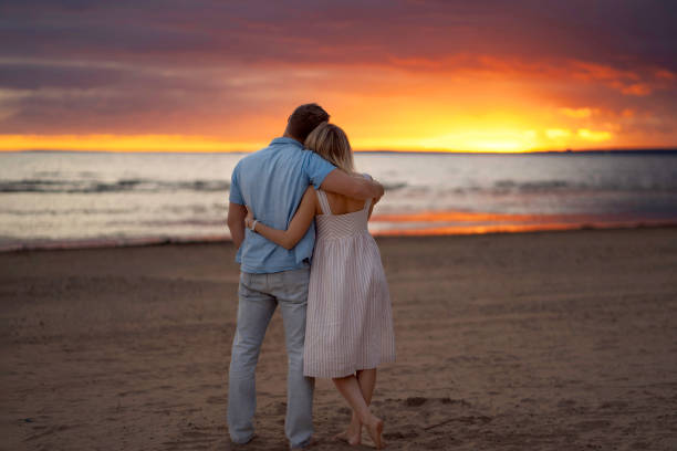 счастливая пара стоит на пляже на закате, обнимается и смотрит на море. beautitul фиолетовое и желтое небо на фоне. романтическое фото для родите - heterosexual пара стоковые фото и изображения