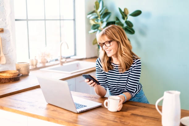 istock Mature woman using her laptop and mobile phone while sitting at the kitchen at home 1333387235
