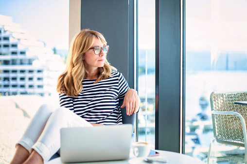 Smiling mature woman sitting on chair and looking thoughtfully while looking out the window at home.