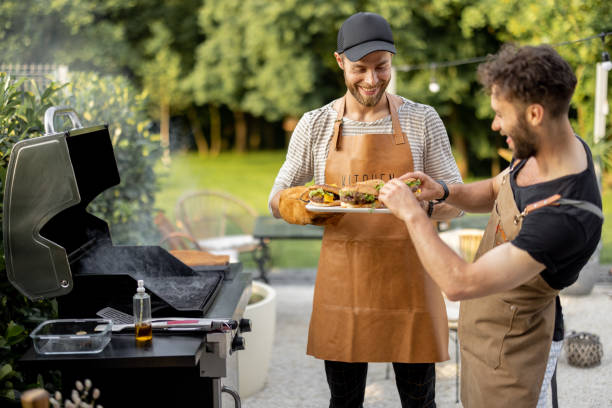 Two guys grilling at backyard Two handsome male friends have fun while grilling meat and buns for burgers on gas grill at backyard on nature grilled stock pictures, royalty-free photos & images