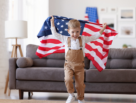 Happy child boy with flag of United States running in living room at home while celebrating independence day of USA on 4th of July with family. American patriotic holiday concept