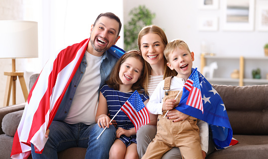 Young happy american family parents and two little kids sitting on sofa at home with flags of united states and smiling at camera while celebrating Independence Day. Patriotic US holiday concept