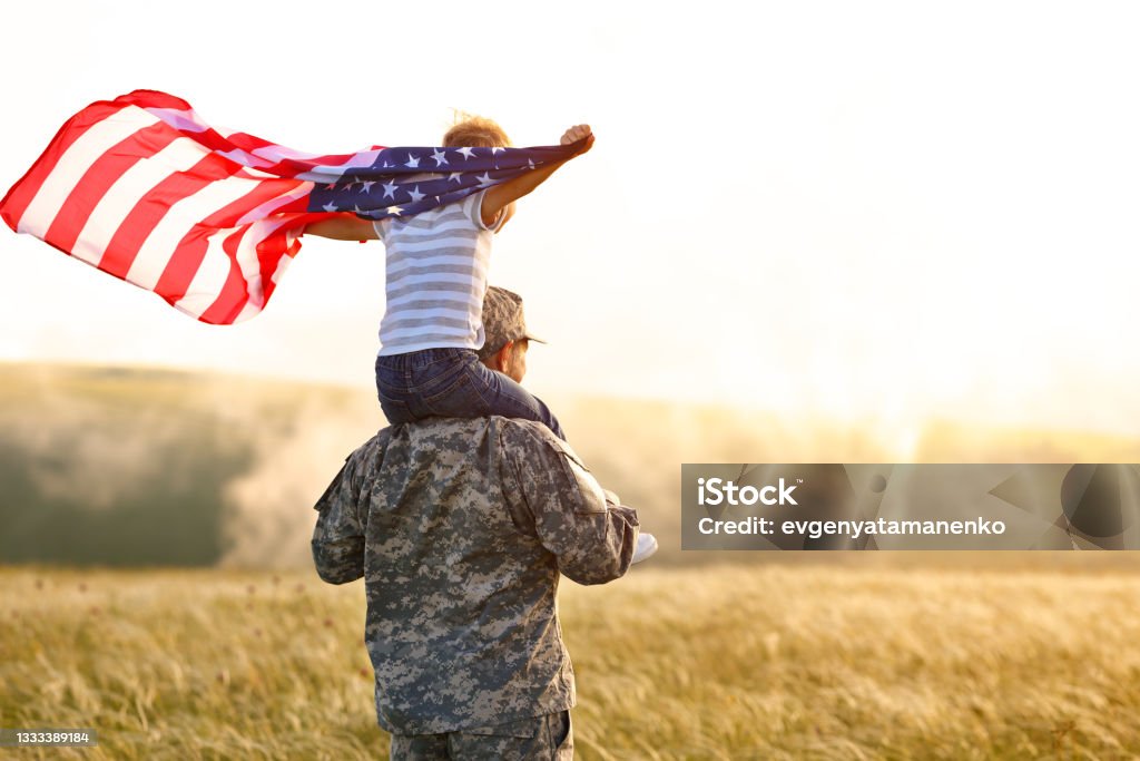 Excited child sitting with american flag on shoulders of father reunited with family Rear view of military man father carrying happy little son with american flag on shoulders and enjoying amazing summer nature view on sunny day, happy male soldier dad reunited with son after US army Veteran Stock Photo