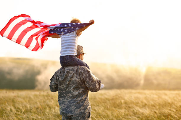 Excited child sitting with american flag on shoulders of father reunited with family Rear view of military man father carrying happy little son with american flag on shoulders and enjoying amazing summer nature view on sunny day, happy male soldier dad reunited with son after US army vet stock pictures, royalty-free photos & images