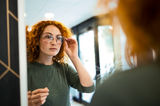 Young woman trying on glasses in optical store looking at mirror