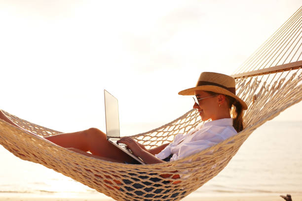 istock Working at sunset. Side view of young happy female using laptop while lying in the hammock on the beach 1333392924