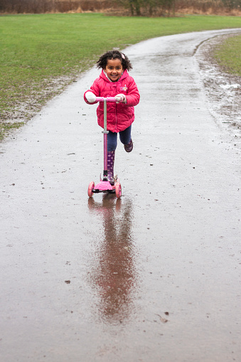 A young mixed race girl wearing a padded raincoat, riding a push scooter on a wet footpath through Plessey woods, northumberland. She is smiling while looking ahead.