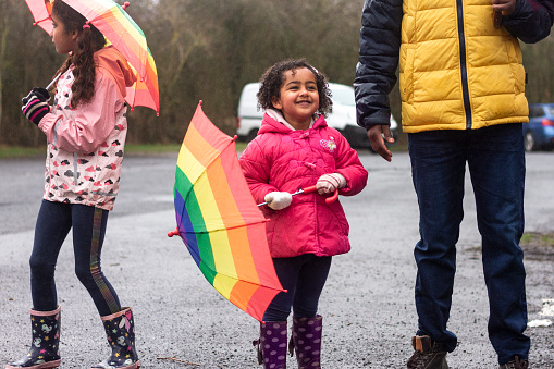 A multi-ethnic family standing in a carpark, getting ready to go on a day trip to Plessey Woods, Northumberland. The main focus is the youngest girl holding an umbrella to her side while the rain falls on her, she is embracing the weather. Her sister and father are standing close by.