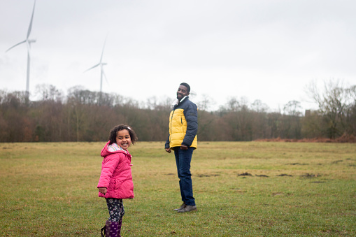 A young girl and her father in a green space in Plessey Woods, Northumberland. The young girl is smiling while looking at the camera and her father is watching her.