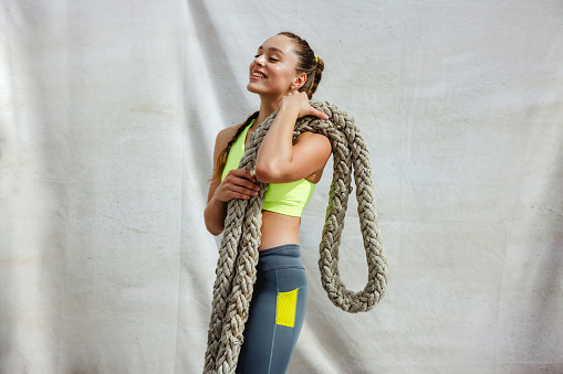 Fit woman in sportswear with battle rope standing on white background. Female athlete relaxing after doing workout routine.