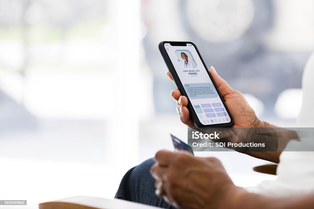 Woman holding credit card selects doctor from tele-medicine site From the comfort of her home, the senior adult woman uses her phone to find a doctor.  She has her credit card ready to pay for the tele-health consultation. Healthcare And Medicine Stock Photo