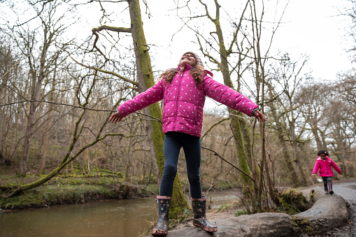 A young girl standing on a log in Plessey Woods, Northumberland. She has her arms outstretched while looking up at the sky and smiling. Her sister is walking along the log in the background.