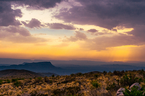 The setting sun shines golden light on the horizon behind storm clouds casting a blue shadow over the landscape.