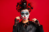 Girl with sugar skull makeup with a wreath of flowers on her head and skull, wearth lace gloves and leather jacket, testing new lip gloss isolated on red background