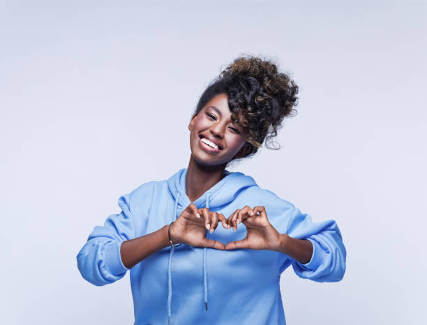 Cheerful young woman showing love sign Portrait of happy african young woman wearing blue hoodie, making heart shape with hands. Studio shot on grey background. i love you photos stock pictures, royalty-free photos & images